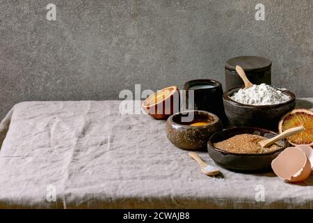 Ingredients for home baking. Flour, eggs, cane sugar, milk in different ceramic bowls, blood oranges and spices on linen table cloth. Grey texture bac Stock Photo