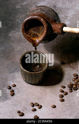 Black turkish coffee pouring from craft handmade ceramic coffee pot cezve with wooden handle into cup, roasted coffee beans above. Grey texture backgr Stock Photo