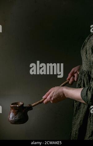 Woman in dark dress hold in hands craft handmade ceramic coffee pot cezve with long wooden handle for making turkish coffee. Stock Photo
