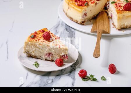 Homemade raspberry baked cheesecake sliced on plate decorated by fresh raspberries, mint, mint and wooden cake server over white marble background. Stock Photo