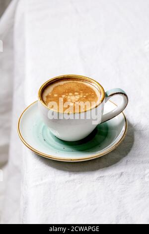 Black coffee espresso with foam in white ceramic cup with saucer standing on white cotton table cloth. Copy space Stock Photo
