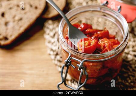 Sun-dried cherry tomatoes olive oil in glass jar with fork, standing on wooden kitchen table with sliced rye bread. Stock Photo
