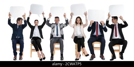 Team of 6 businessmen holding blank speech bubble above their heads, one hiding his face while sitting on chairs on white studio background Stock Photo