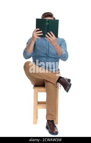 young casual man covering face with book and reading, sitting isolated on white background, full body Stock Photo