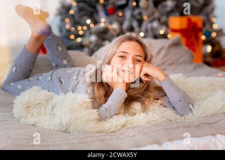 Dreamful cute teen girl near Christmas tree. Kid wearing pajama dreaming near tree in the morning. Merry Christmas and Happy Holidays concept Stock Photo