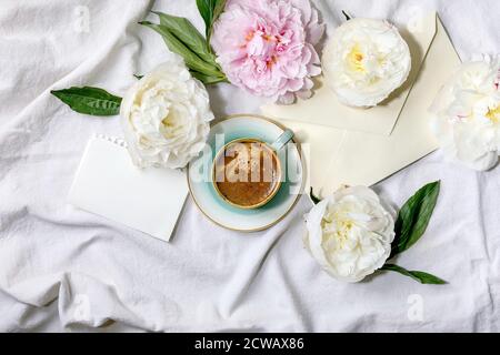 Cup of espresso coffee, blank paper, envelope, pink and white peonies flowers with leaves over white cotton textile background. Flat lay, copy space Stock Photo