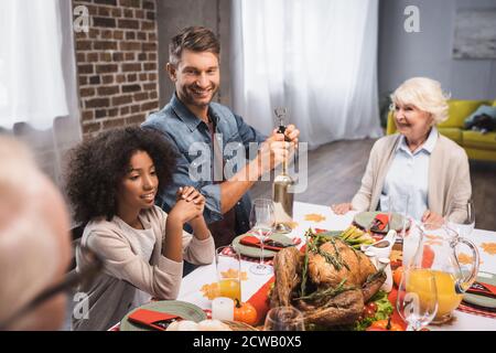 selective focus of man opening bottle of white wine during thanksgiving dinner with multicultural family Stock Photo