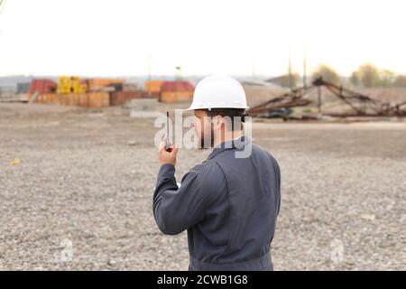 Back view of male foreman standing on construction site, talking by VHF walkie talkie and holding blueprints. Stock Photo