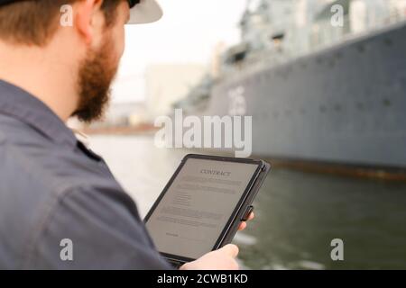 Navigation officer reading contract on tablet near vessel in background. Stock Photo