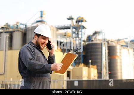 Industrial euroepean engineer talking by VHF walkie talkie and holding papers near factory outside. Stock Photo