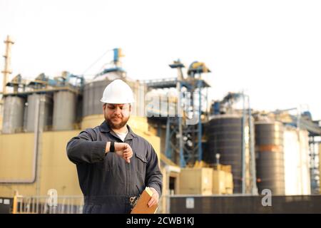 Supervisor looking at watch and holding papers notes near factory outside. Stock Photo