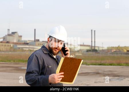 Portrait of male foreman standing on construction site, talking by walkie talkie and holding papers. Stock Photo