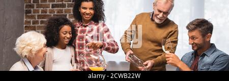 panoramic shot of senior man and african american woman pouring orange juice and white wine during thanksgiving dinner with multicultural family Stock Photo