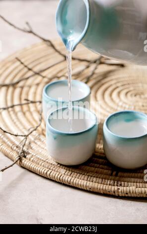 Sake ceramic set for traditional japanese alcohol drink rice wine sake pouring from pitcher in three cups, standing on straw napkin with dry branches Stock Photo