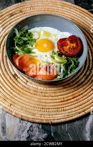 Healthy breakfast bowl with fried eggs, salmon, avocado, grilled tomato and salad serving with bread on straw napkin over dark marble background. Stock Photo