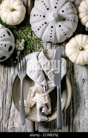 Holiday table setting decoration with white decorative pumpkins, craft clay pumpkins, thuja branches, empty plate with cloth napkin, cutlery over old Stock Photo