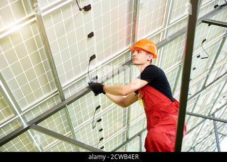 Close up of male is inspecting quality solar panels. Worker in orange uniform and helmet with equipment. Photovoltaic power supply systems. Green energy concept design. Stock Photo
