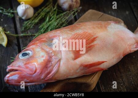 Raw fish grouper on wooden cutting board with sliced lemon, rosemary and saltover old wooden kitchen table. Dark rustic style. Stock Photo