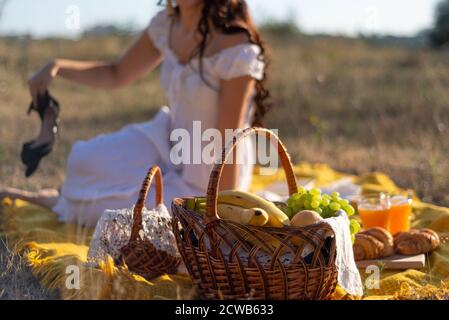 girl in a hat and white dress sitting on a picnic in a grass field in defocus blur. On the cross plan are baskets with fruit. Rustic style, against th Stock Photo