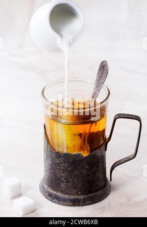 Milk pouring from white jug to retro cup of hot tea in vintage glass-holder with tea spoon and sugar cubes over white marble backgtound. Stock Photo