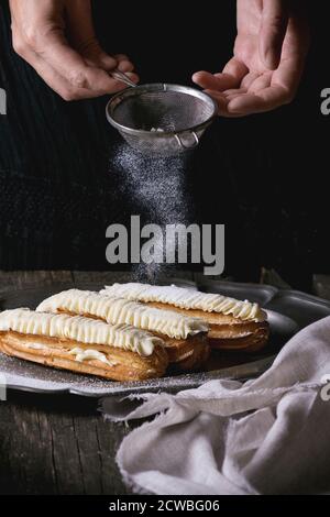 Eclairs with white butter cream in vintage metal plate with Sifting sugar powder from woman's hands. Dark rustic style. Day light. Outside view Stock Photo