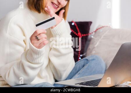 A young woman in a white sweater, Santa Claus hat makes online purchases using a notebook and a credit card. New year shopping and christmas sale Stock Photo