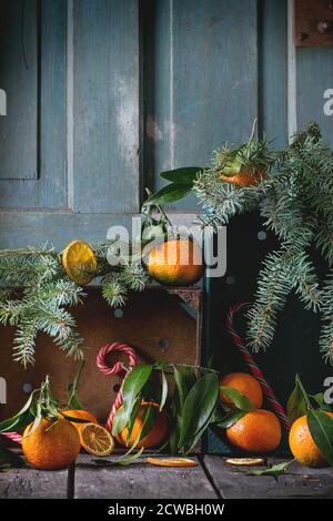 Tangerines with leaves in Christmas decor with Christmas tree, dry orange and candies in wooden boxes over old wooden table. Dark rustic style Stock Photo