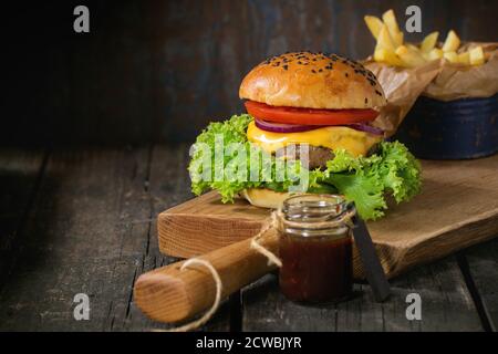 Fresh homemade burger with black sesame seeds on wooden cutting board with fried potatoes, served with ketchup sauce in glass jar over old wooden tabl Stock Photo