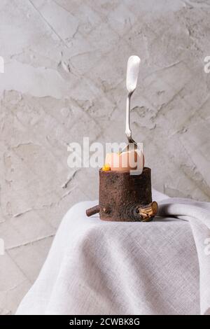 Breakfast with started eating soft-boiled egg with pouring yolk in wooden eggcup with silver spoon on white cloth over white table. With plastered wal Stock Photo