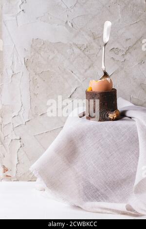 Breakfast with started eating soft-boiled egg with pouring yolk in wooden eggcup with silver spoon on white cloth over white table. With plastered wal Stock Photo