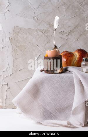 Breakfast with started eating soft-boiled egg with pouring yolk in wooden eggcup and home made bread served with salt and silver spoon on wooden chest Stock Photo