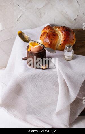 Breakfast with started eating soft-boiled egg with pouring yolk in wooden eggcup and bread served with salt and silver spoon on wooden chest with whit Stock Photo