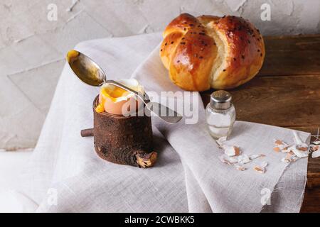 Breakfast with started eating soft-boiled egg with pouring yolk in wooden eggcup and home made bread served with salt and silver spoon on wooden chest Stock Photo