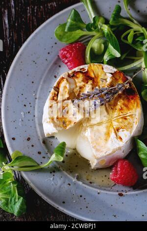 Melting sliced grilled goat cheese on blue plate, served with liquid honey, lavender, raspberries, wholegrain toast and green salad. Over old wooden t Stock Photo