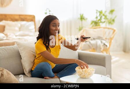 Young African American woman sitting on couch, eating popcorn and watching interesting show on television, indoors Stock Photo