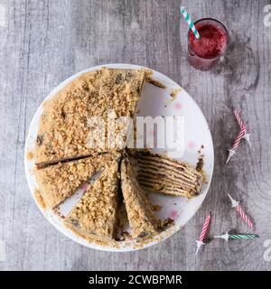 Sliced Homemade Birthday Honey Cake with chocolate cream, served on white ceramic plate over white wooden table with b-day candles and glass of red ju Stock Photo