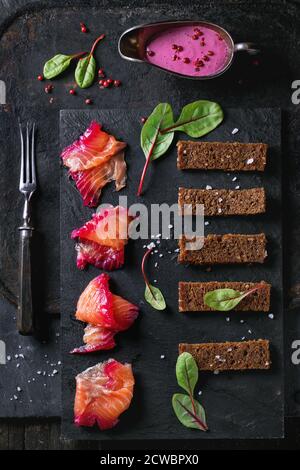 Sliced salmon filet, salted with beetroot juice, served with whole wheat toasts, salad leaves, beetroot sauce and pink pepper over black slate surface Stock Photo