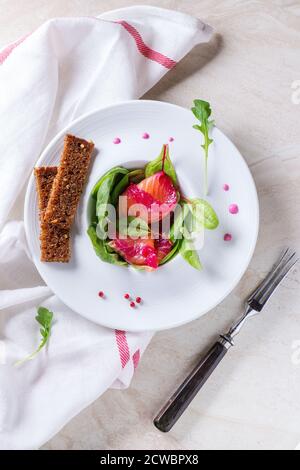 White plate with Sliced salmon filet, salted with beetroot juice, served with whole wheat toasts, salad leaves and beetroot sauce over white marble su Stock Photo