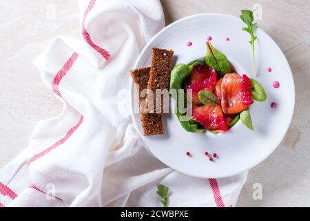 White ceramic plate with Sliced salmon filet, salted with beetroot juice, served with whole wheat toasts, salad leaves and beetroot sauce over white m Stock Photo
