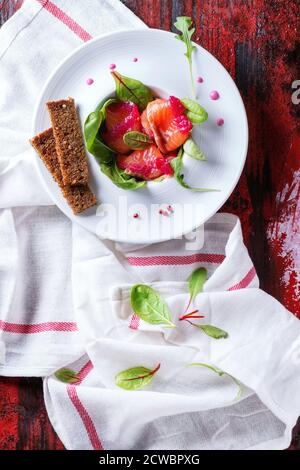 White ceramic plate with Sliced salmon filet, salted with beetroot juice, served with whole wheat toasts, salad leaves and beetroot sauce over red woo Stock Photo