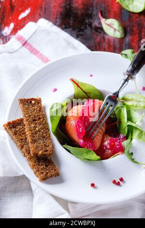 White ceramic plate with Sliced salmon filet, salted with beetroot juice, served with whole wheat toasts, salad leaves and beetroot sauce over red woo Stock Photo