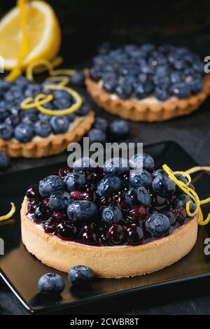 Lemon Tart and tartlets with fresh and cooked blueberries, served on black square plate with lemon and lemon zest over black background. Stock Photo