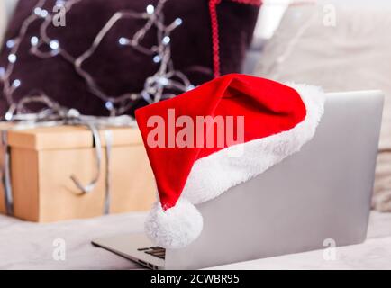 Christmas laptop in santa hat standing on a sofa, a free space. Remote work on Christmas Eve. Online store, online ordering gifts for the holidays. Stock Photo