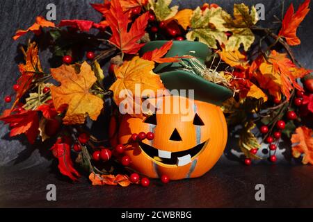 Ceramic Orange Halloween Pumpkin with Smile and Green Hat. Artificial Autumn Leaves. Halloween and Fall Vibes. Stock Photo