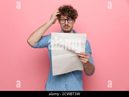 shocked young guy reading newspaper, holding hand to head and having a headache, standing on pink background Stock Photo