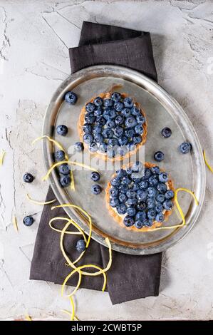 Two Lemon tartlets with fresh blueberries, served on vintage metal tray with lemon zest and black textile napkin over gray plastered surface. Flat lay Stock Photo