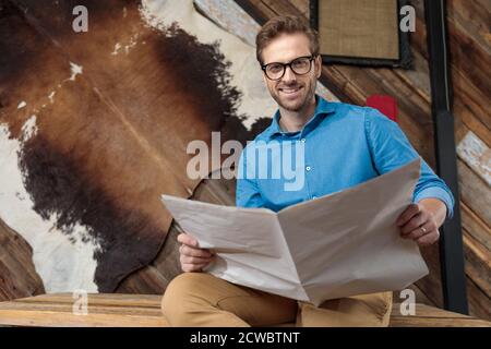 Positive model smiling and reading a newspaper while wearing blue shirt and glasses, sitting on a desk on coffeeshop background Stock Photo
