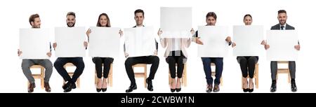 Team of 8 businessmen holding empty billboards while one is hiding her face while sitting on chairs on white studio background Stock Photo