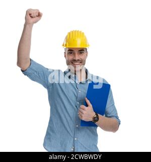 Cheerful casual man celebrating and holding clipboard, wearing safety helmet, standing on white studio background Stock Photo