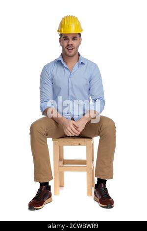 Confused casual man frowning and solving problem, wearing hard hat while sitting on a chair on white studio background Stock Photo